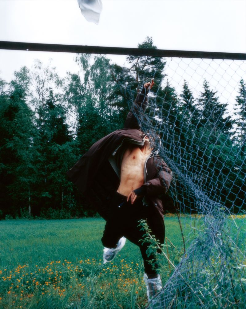 A man leaps dynamically outdoors behind a wire mesh fence, wearing an open dark brown jacket revealing his torso over black pants.