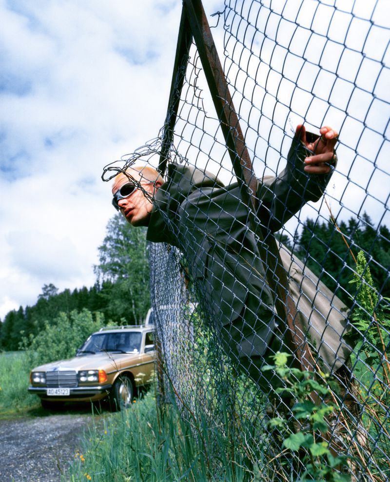 Man in sunglasses climbs through damaged chain-link fence outdoors during daytime; classic car and trees in background; eye-level angle.