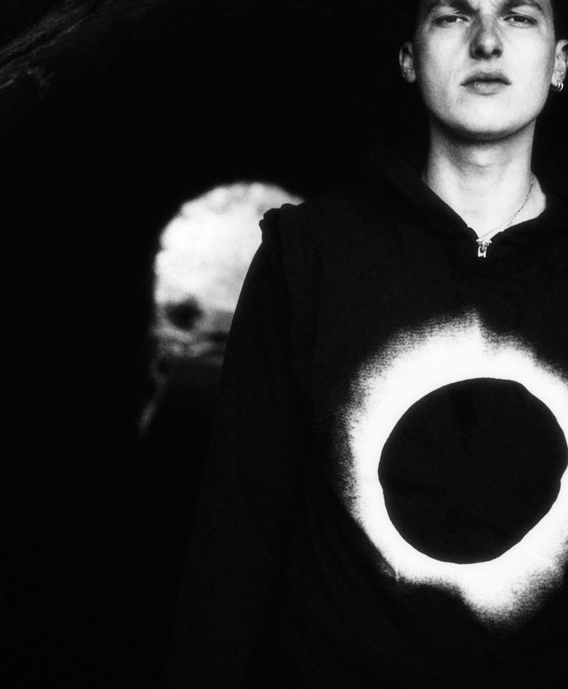 Monochrome portrait of a young adult with fair skin wearing a black hoodie with a circular graphic, against a blurred background.