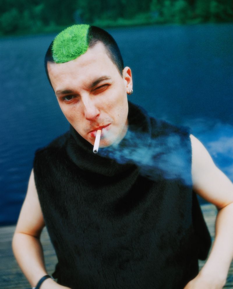 Portrait of a young adult male smoking a cigarette outdoors, sporting a green mohawk, dark vest, winking and blowing smoke.