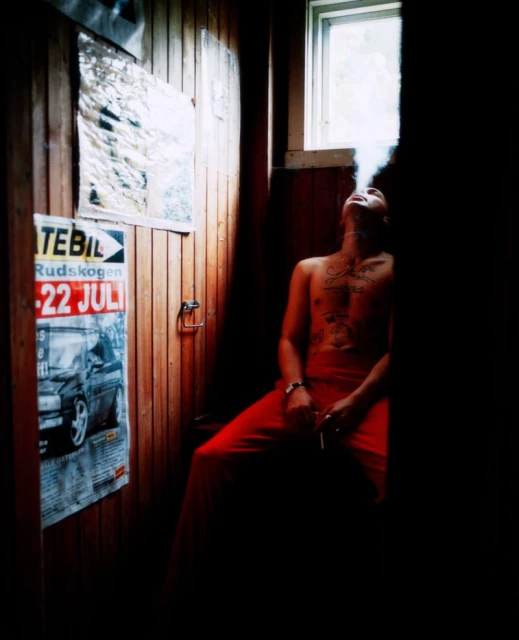 Tattooed man in red pants exhales smoke in an indoor wood-paneled room with window light and aged posters, capturing a dimly lit, casual scene.