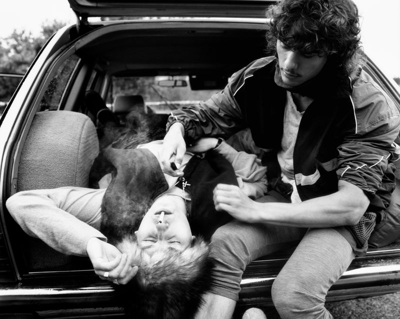 Black and white image of two men in a car, one seated and one reclining, with cigarette smoke in the foreground and neutral background.