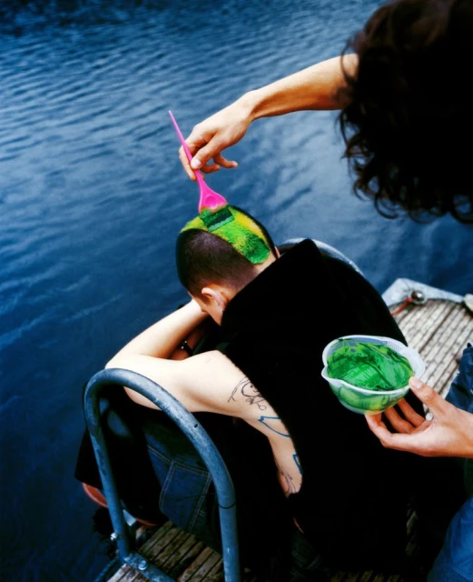 Person getting their head dyed green with a brush and bowl of green dye outdoors near the water on a sunny day. 
