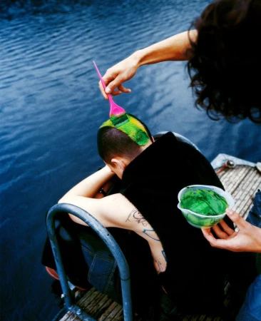 Person getting their head dyed green with a brush and bowl of green dye outdoors near the water on a sunny day. 
