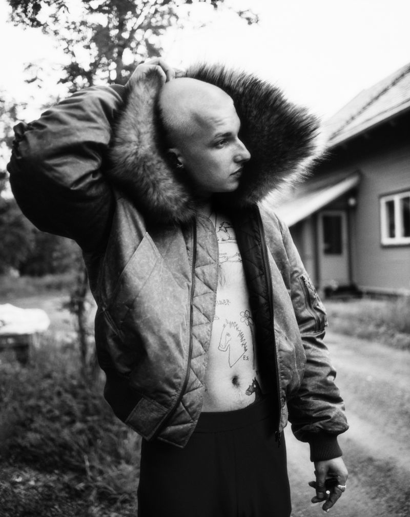 Monochrome portrait of a bald person with tattoos, wearing a fur-lined jacket, standing outdoors against a blurred background of a building.