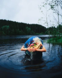 A person wearing a blue patterned wrap leans over dark lake water, splashing water onto their face in front of a pine forest.
