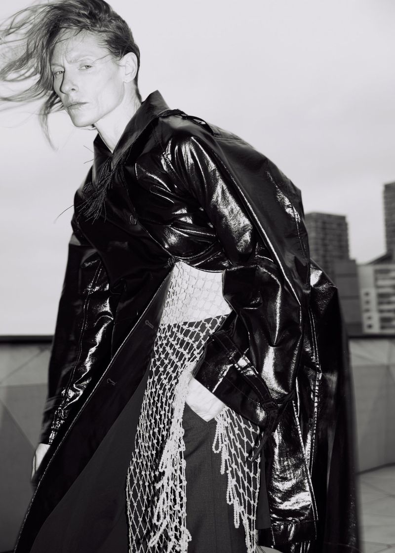 Monochrome portrait of adult female fashion model in shiny black trench coat with netting detail, city buildings in the background.