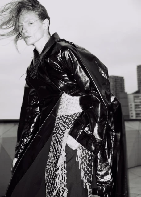 Monochrome portrait of adult female fashion model in shiny black trench coat with netting detail, city buildings in the background.