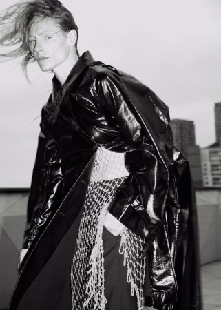 Monochrome portrait of adult female fashion model in shiny black trench coat with netting detail, city buildings in the background.
