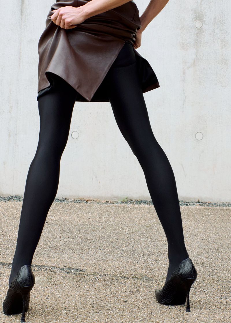 Fashion shot of a woman posing in a brown leather dress, black tights, and black high heels against a concrete background, legs visible.
