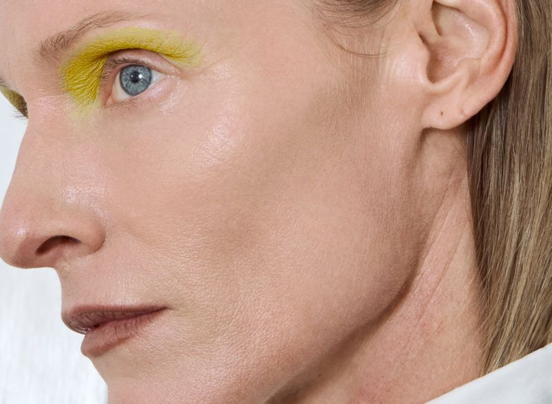 Close-up portrait of an adult female with fair skin, light blue eyes, and yellow eyeshadow, against a white background.
