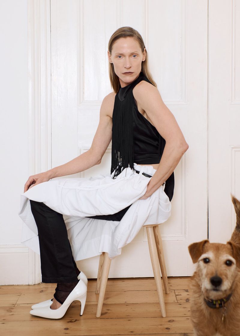 A slender adult woman in a black top, white skirt, and heels is sitting on a wooden stool in a minimalist room with a brown dog.