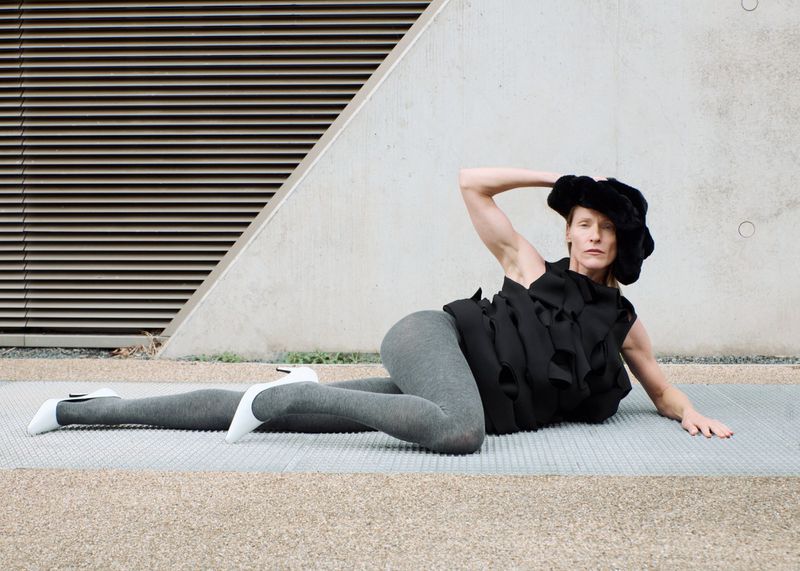 Adult woman poses on a grey tiled surface wearing a black textured top, grey leggings, and white heels, set against a geometric concrete backdrop.