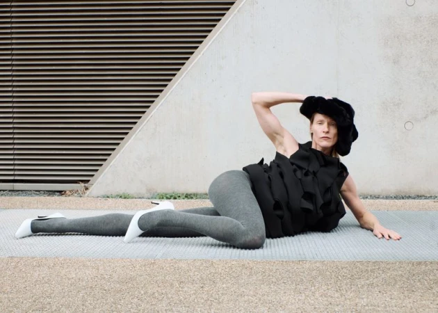 Adult woman poses on a grey tiled surface wearing a black textured top, grey leggings, and white heels, set against a geometric concrete backdrop.
