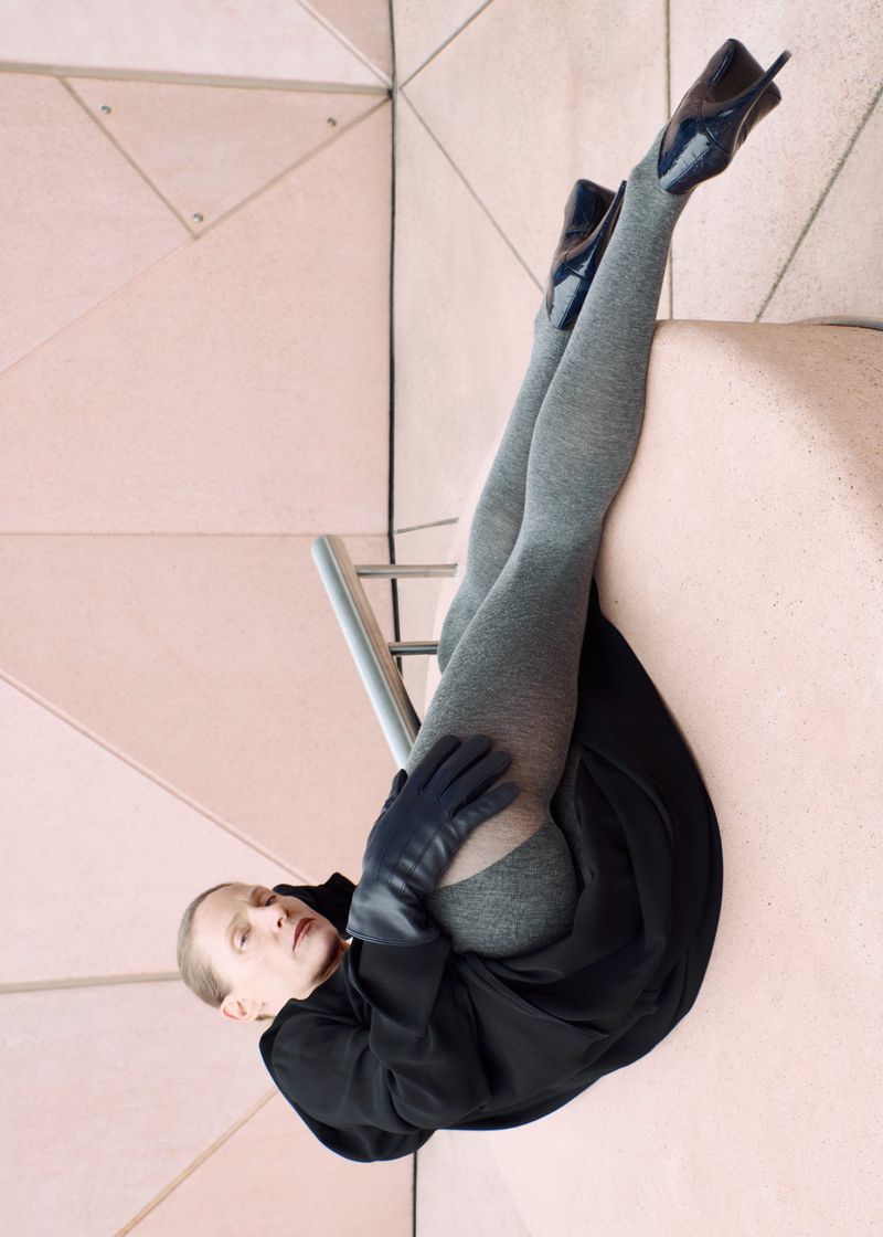 Fashion image features a model in black coat, grey tights, and high heels posing against a geometric pale pink tiled background.