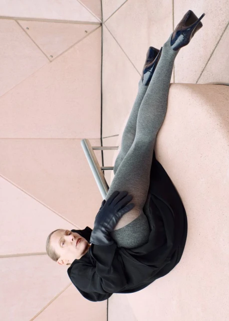 Fashion image features a model in black coat, grey tights, and high heels posing against a geometric pale pink tiled background.