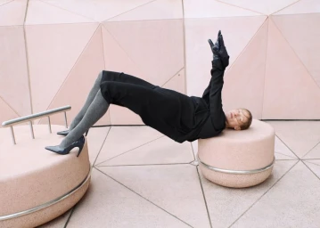 Model Lindsay Sullivan posing in a bridge pose across two pink stone pedestals wearing a black outfit, grey tights, and pointed heels.