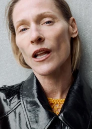 Lindsay Sullivan wearing a glossy black jacket and yellow beaded necklace against a textured grey wall.