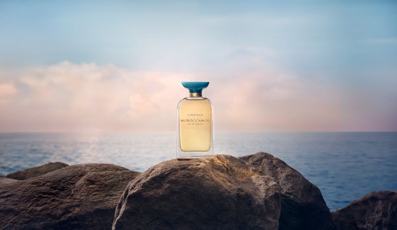 Glass perfume bottle with a blue cap sits on a rocky surface with the ocean and sky visible in the blurred background.