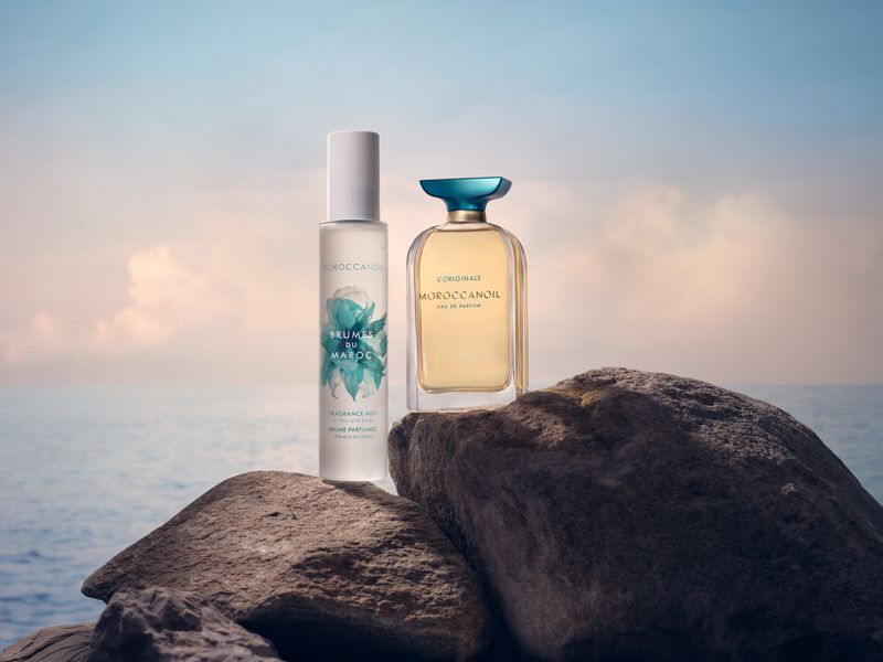 Moroccanoil fragrance mist and Eau de Parfum bottles displayed on textured rock surface with a blurred ocean and sky background.