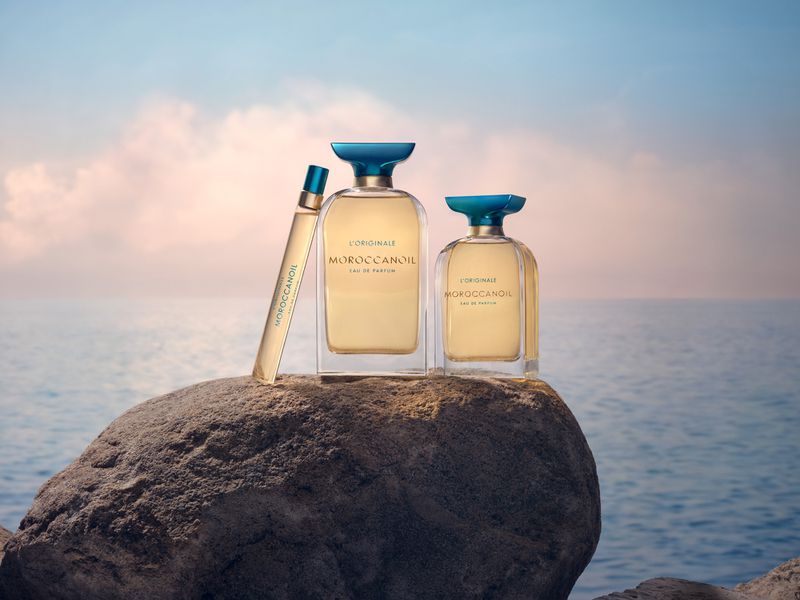 Three Moroccanoil perfume bottles displayed on a rock against a blurred background of ocean and sky, showcasing beauty and fragrance.
