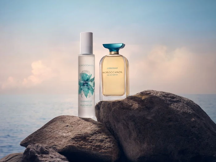 Two perfume bottles resting on dark rocks against an ocean and sunset sky background.