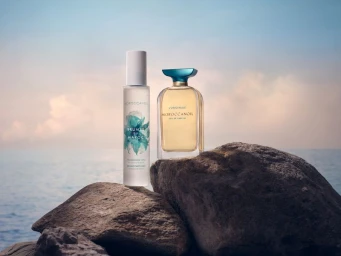 Two perfume bottles resting on dark rocks against an ocean and sunset sky background.