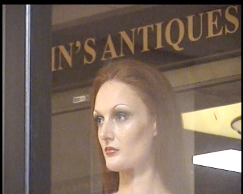 A mannequin with auburn hair and makeup stands inside an antique store, as seen through the glass storefront window.