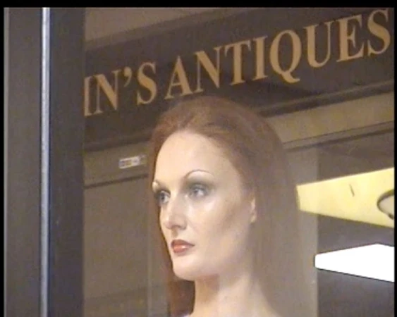 A mannequin with auburn hair and makeup stands inside an antique store, as seen through the glass storefront window.