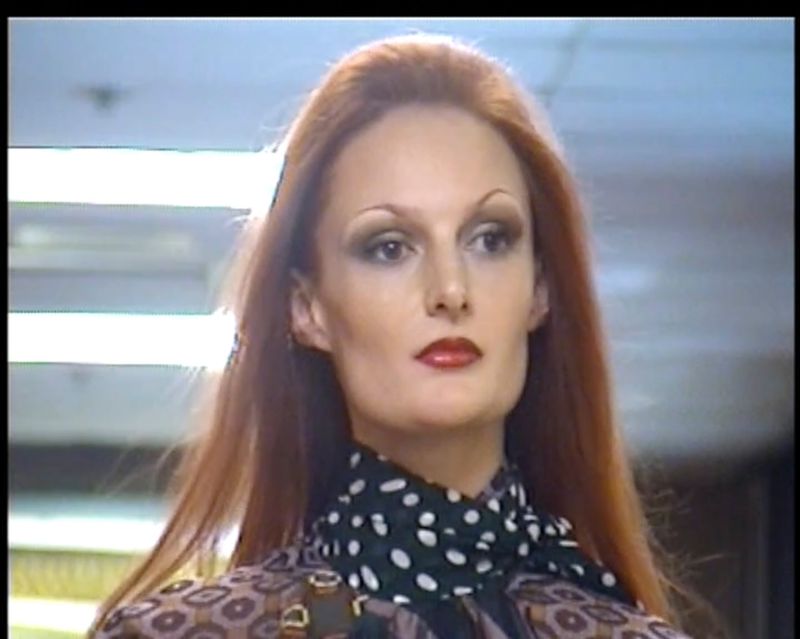 Portrait of a red-haired woman with makeup, wearing a polka dot scarf and patterned jacket, in front of a blurred office background.