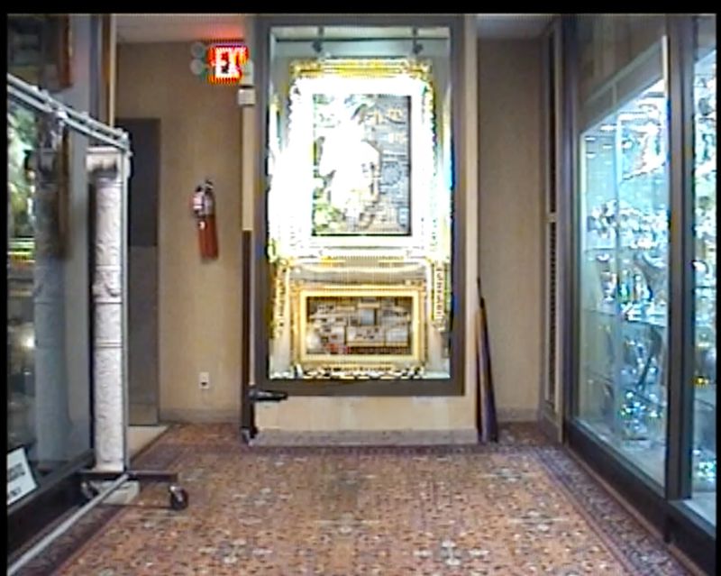 A collection of framed artwork is displayed inside glass cabinets in an indoor museum setting, with an exit sign visible in the background.