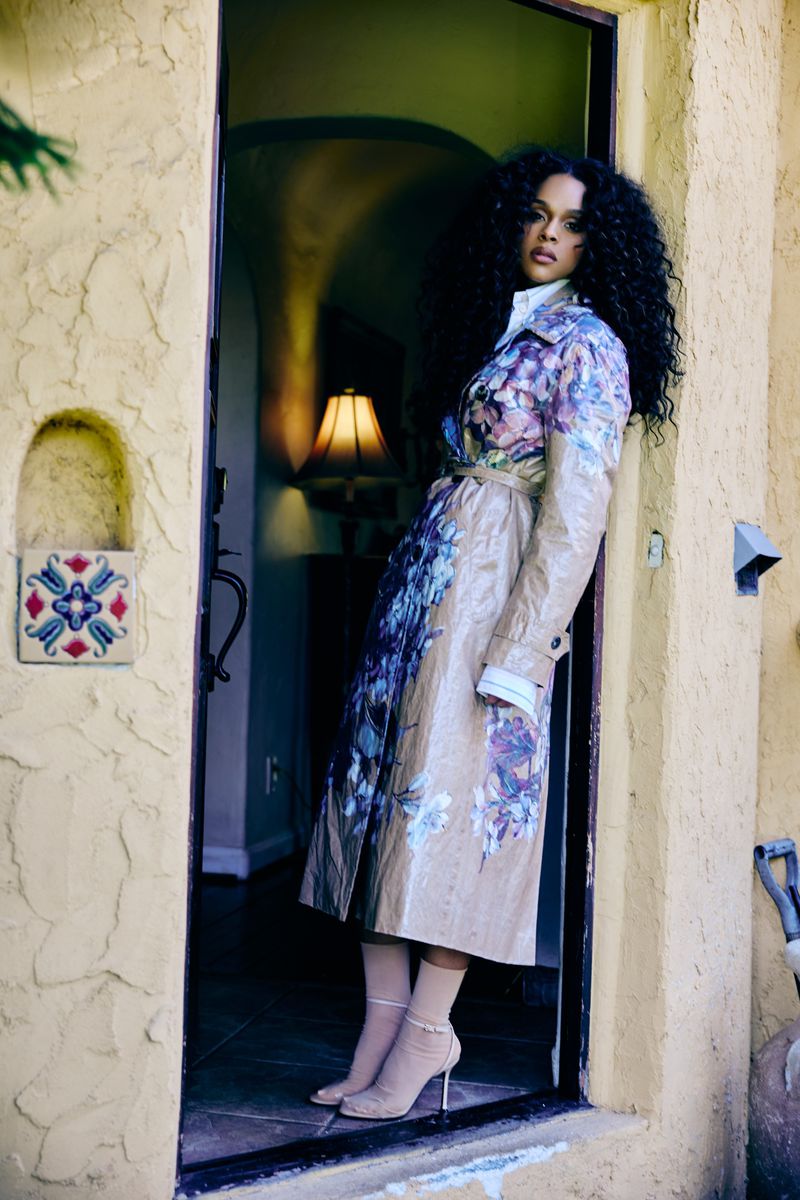 Woman in a floral trench coat poses in a doorway with visible curly hair, pantyhose, and heels. The scene includes a lamp and outdoor greenery.