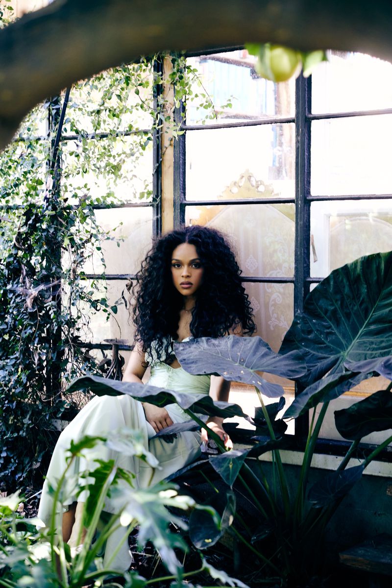 Portrait of woman with curly hair in light dress sitting amid lush green foliage in sunlit garden, greenhouse setting with large windows.