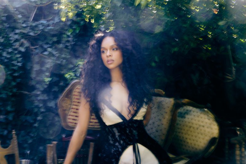 Woman with curly hair wearing a low-cut black and cream top poses outdoors among greenery and antique chairs in soft, romantic lighting.