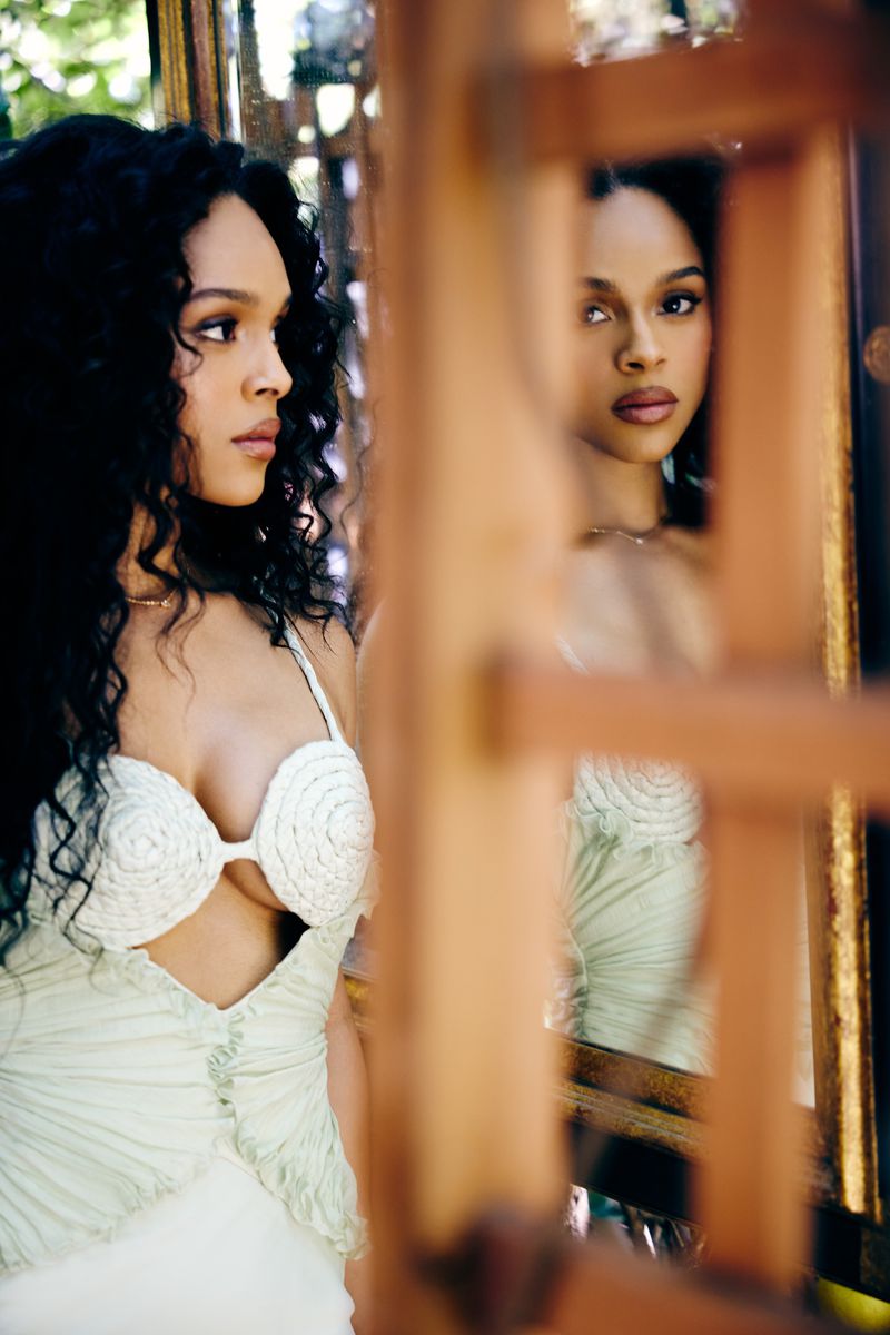 Elegant woman in a pale gown looks pensively to the side, reflected in a vintage mirror amid dappled light and natural greenery.
