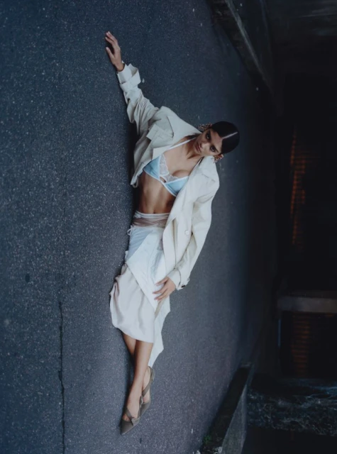 Fashion shot of a stylish adult woman in a light-colored coat, lace bra, and skirt lying on a dark asphalt road in an urban setting.