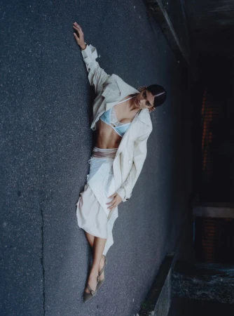 Fashion shot of a stylish adult woman in a light-colored coat, lace bra, and skirt lying on a dark asphalt road in an urban setting.
