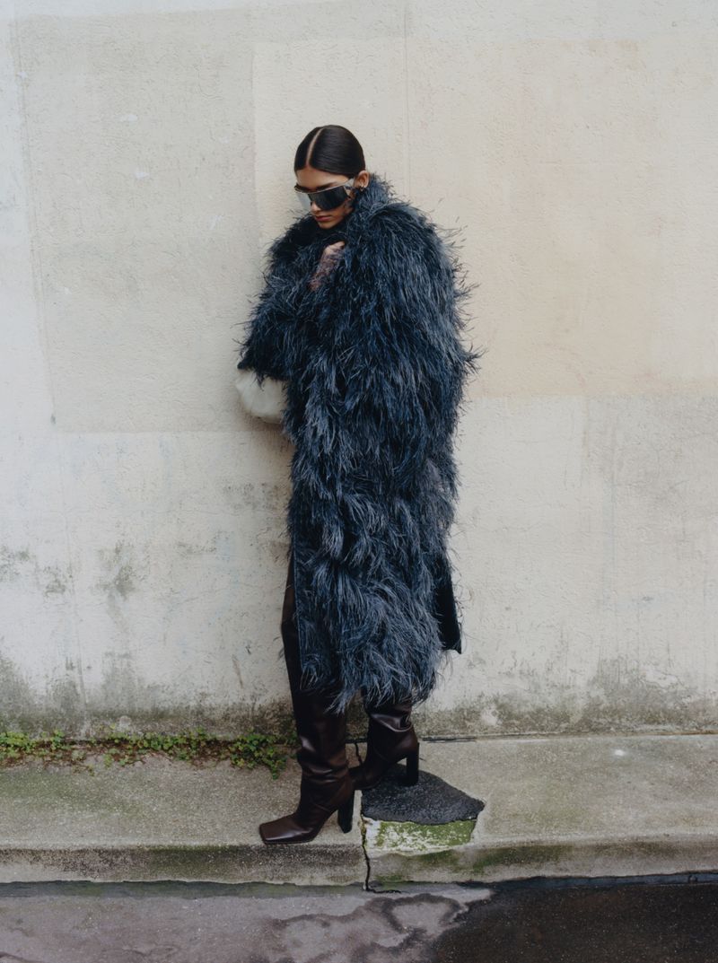 Woman in a feathered coat and boots stands in an urban setting against a wall, with a wet street nearby, posing for a fashion shot.
