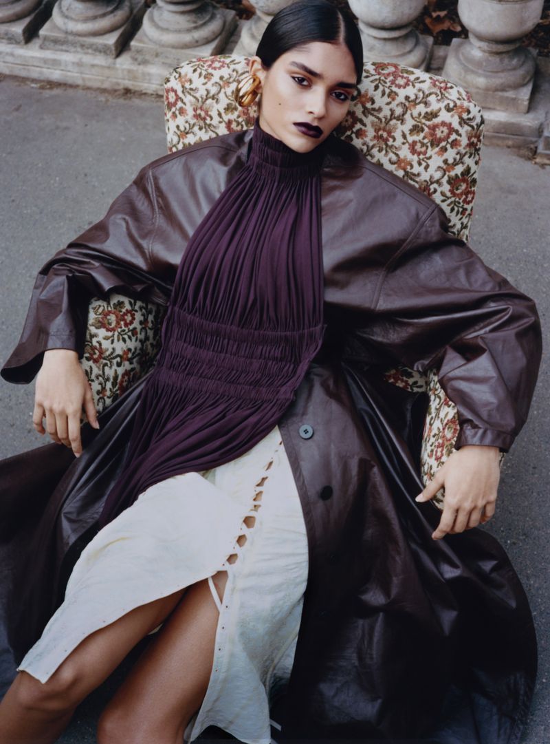 Woman with dark lipstick and brown coat poses in a vintage armchair in an outdoor setting with stone columns in the background.