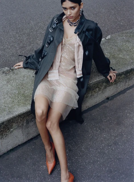 Woman in olive trench coat and nude dress sitting outdoors, orange heels, medium shot, fashion editorial, diffused lighting.