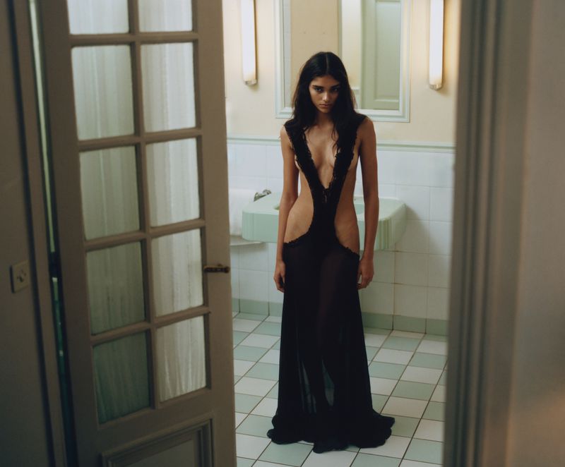Brunette woman in a long sheer black dress with cutouts posing in a bathroom with tiled floors and white walls and fixtures.