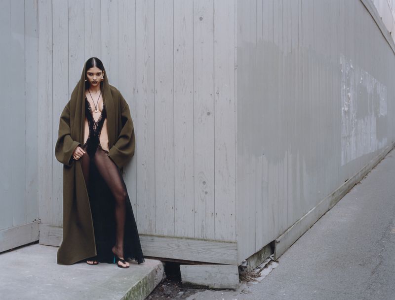 Fashion editorial shot of a woman in a black lace bodysuit, coat and sheer tights, posing against a gray concrete wall in an urban setting.