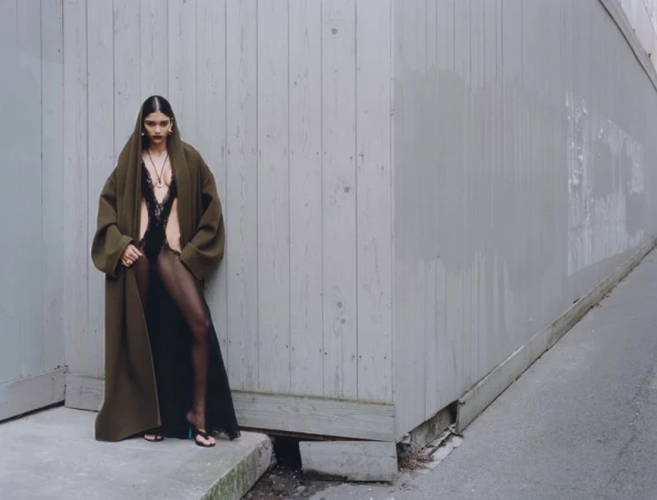 Fashion editorial shot of a woman in a black lace bodysuit, coat and sheer tights, posing against a gray concrete wall in an urban setting.