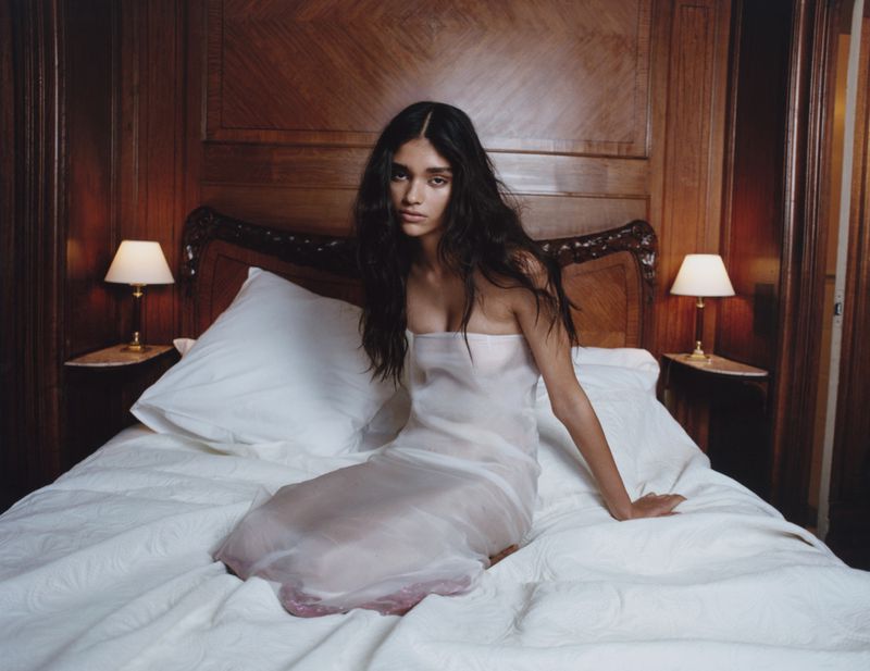 Woman in white dress sitting on a bed in an indoor bedroom with white bedding, lamps on bedside tables, and a dark wood headboard.