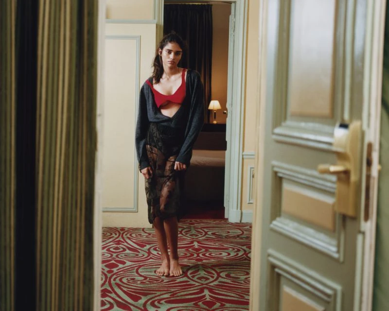 Young woman in lingerie and sheer dress stands in a hotel room doorway with a patterned carpet and muted neutral walls.