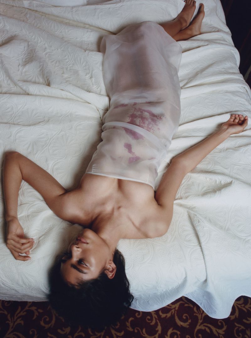 Woman in sheer dress lies on patterned bed. Dark hair frames her face as she extends arms overhead in indoor setting.