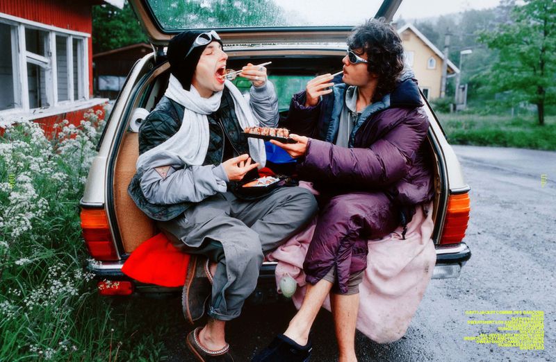 Two young adults in puffer jackets sit in a car hatchback eating sushi in a rural setting, captured in a candid, casual lifestyle scene.