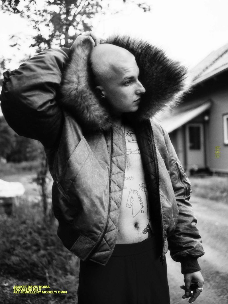 Monochrome portrait of a bald adult male in a fur-collared jacket standing outdoors, looking to the side with a neutral expression.