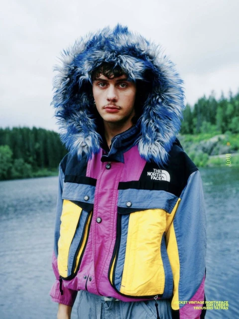 A young adult man poses outdoors in a vibrant, multicolored North Face hooded jacket with a fur-trimmed hood, set against a lake.