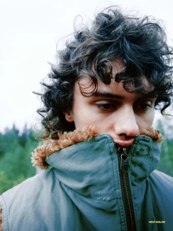 Close-up portrait of a man with dark curly hair wearing a teal shearling-lined high-collar vest outdoors.